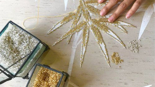 Handmade gold and white embroidered fabric with a person's hands and small containers of gold thread.